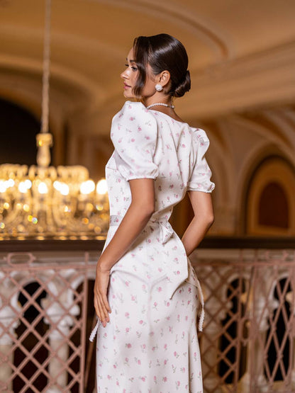 Woman in a white dress with floral patterns standing in an elegant interior setting.