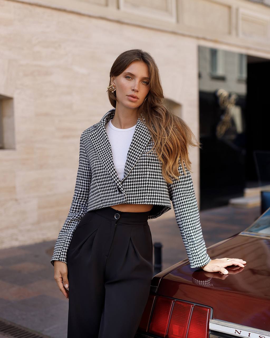 Woman in a checkered jacket and black pants standing next to a red car with a building in the background