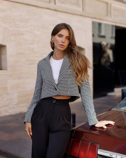 Woman in a checkered jacket and black pants standing next to a red car with a building in the background