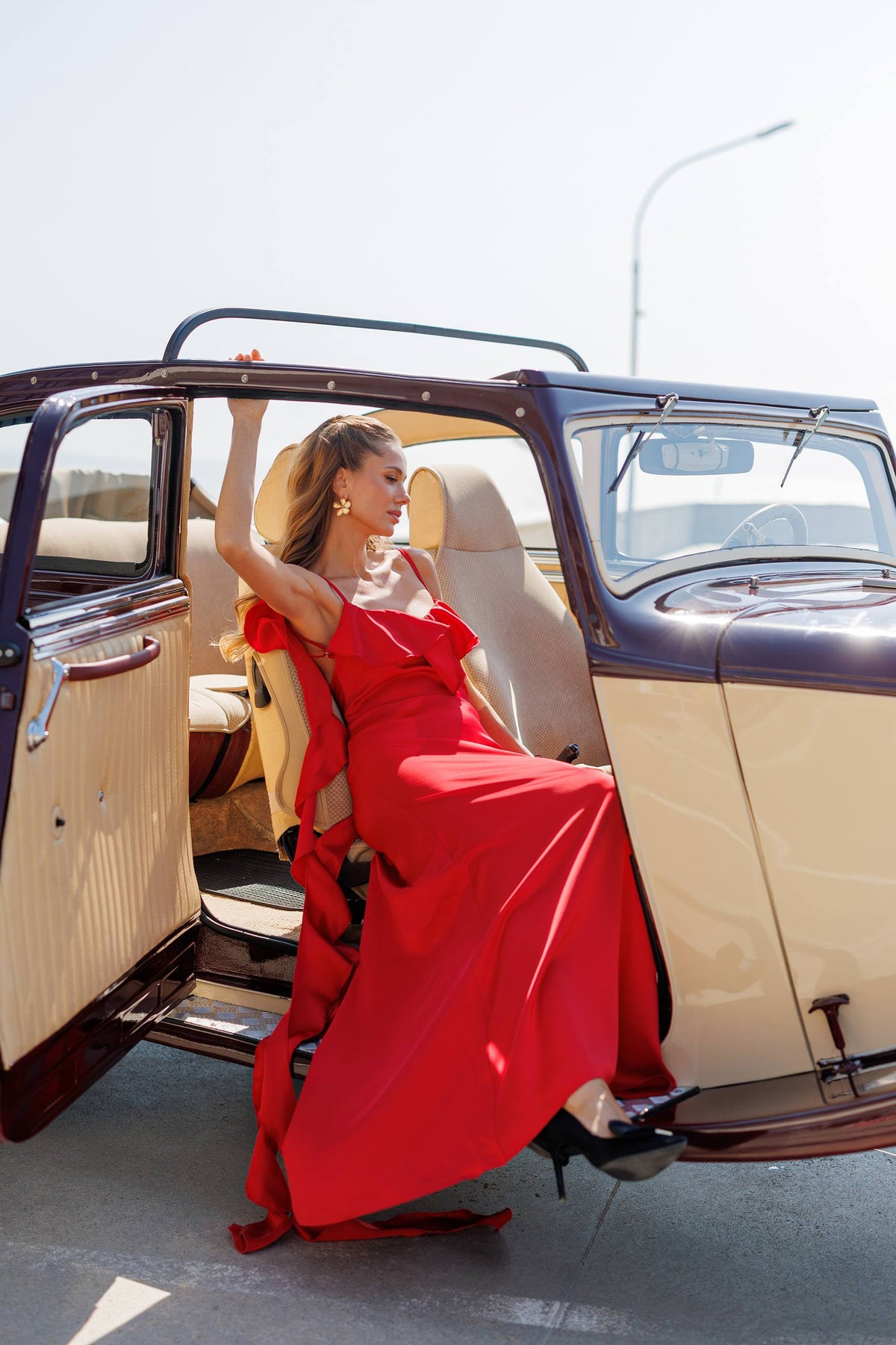 A woman in a flowing red evening gown sits in the open rear door of a vintage cream and dark brown car outdoors on a sunny day.