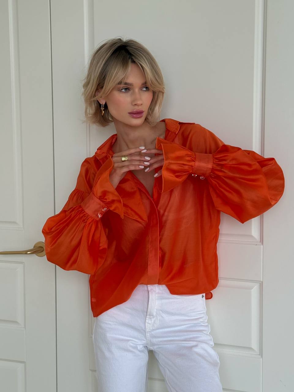 Woman wearing an orange blouse with puff sleeves and white pants against a white door.