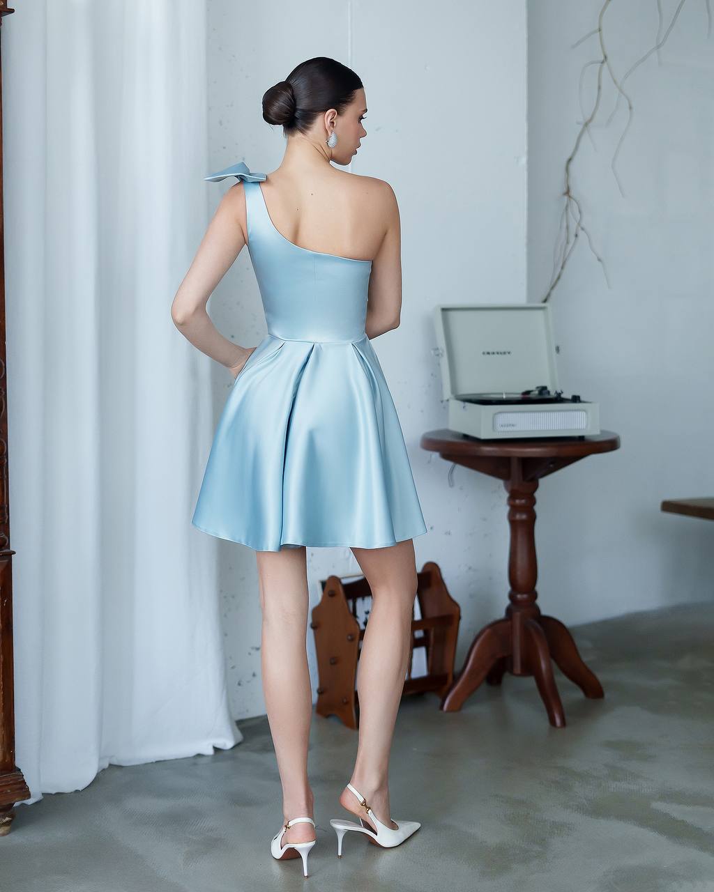 Woman in a light blue one-shoulder dress standing in a room with a white curtain and wooden table.