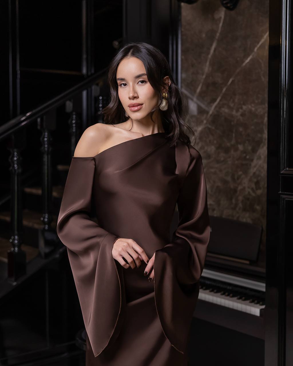 Woman in a brown off-shoulder dress standing in an elegant interior setting.