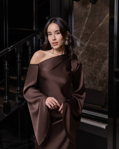 Woman in a brown off-shoulder dress standing in an elegant interior setting.