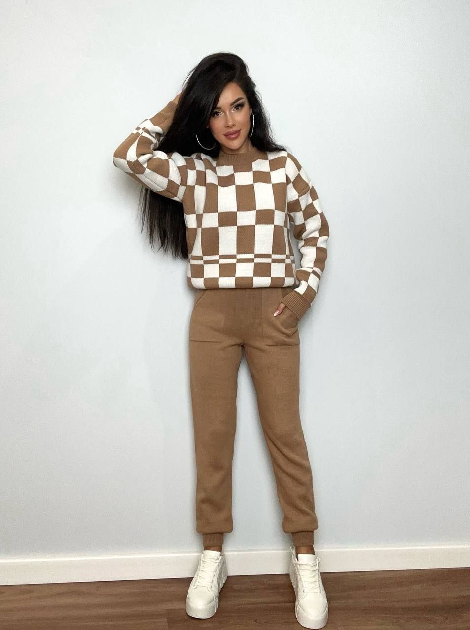 Woman wearing a checkered sweater and brown pants against a white wall.