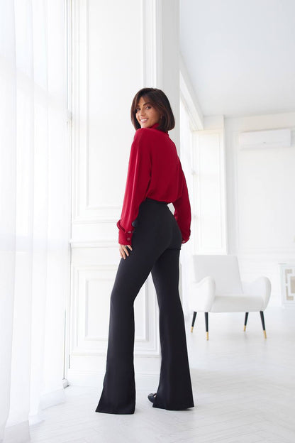 Woman wearing a red blouse and black pants in a bright room.