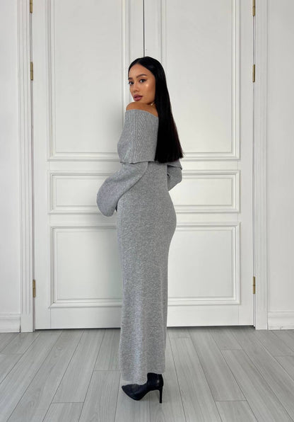 Woman wearing a gray off-shoulder dress standing against a white paneled wall.