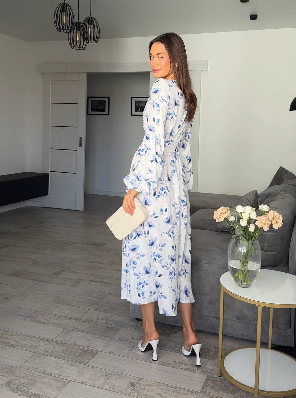 Woman in a floral dress standing in a living room with a gray sofa and a small table with flowers.