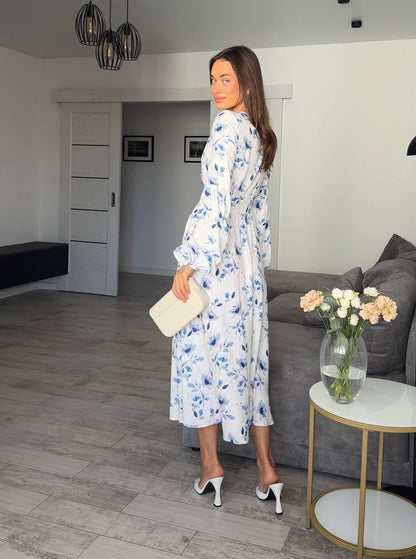Woman in a floral dress standing in a living room with a gray sofa and a small table with flowers.