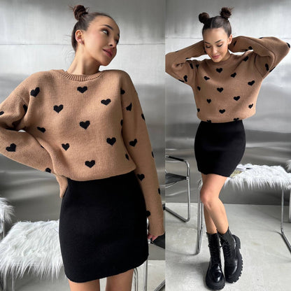 Woman wearing a beige sweater with black heart patterns and a black skirt indoors.