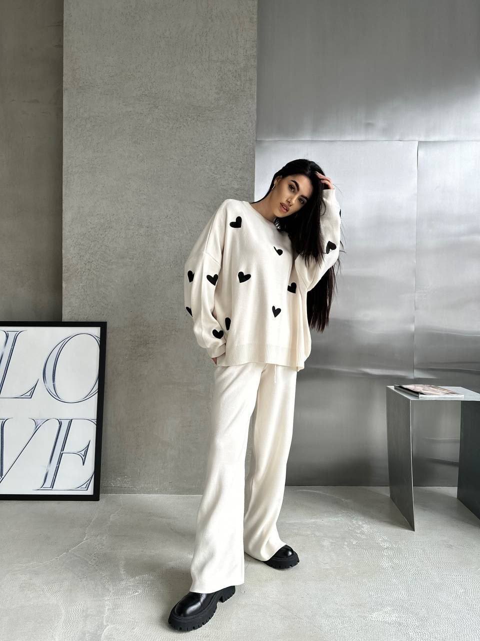 Woman in a white outfit with black heart patterns standing in a modern interior setting.