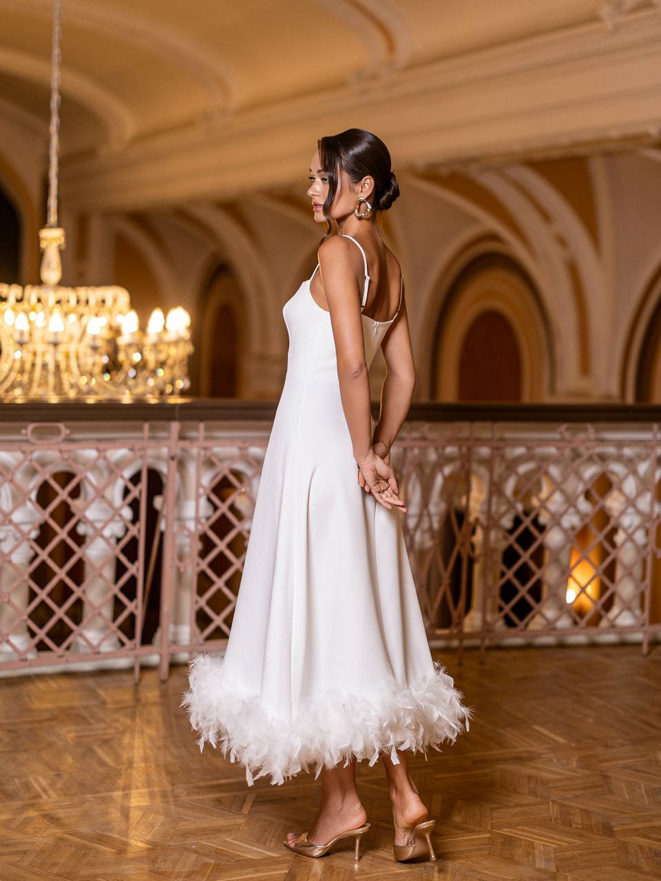 Woman in a white dress with feather details standing in an elegant indoor setting.
