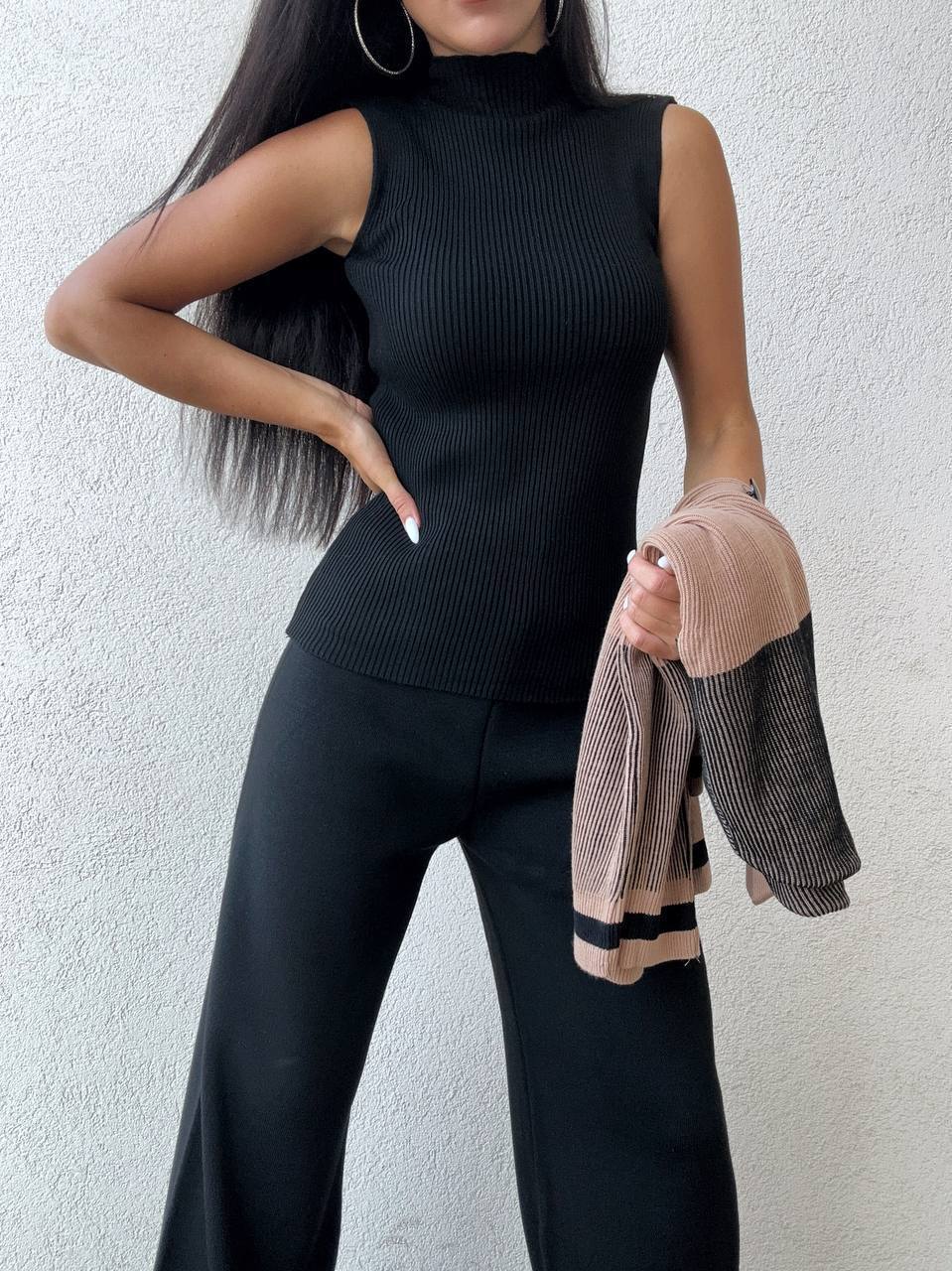 Person wearing a black sleeveless top and pants holding a pink jacket against a white background