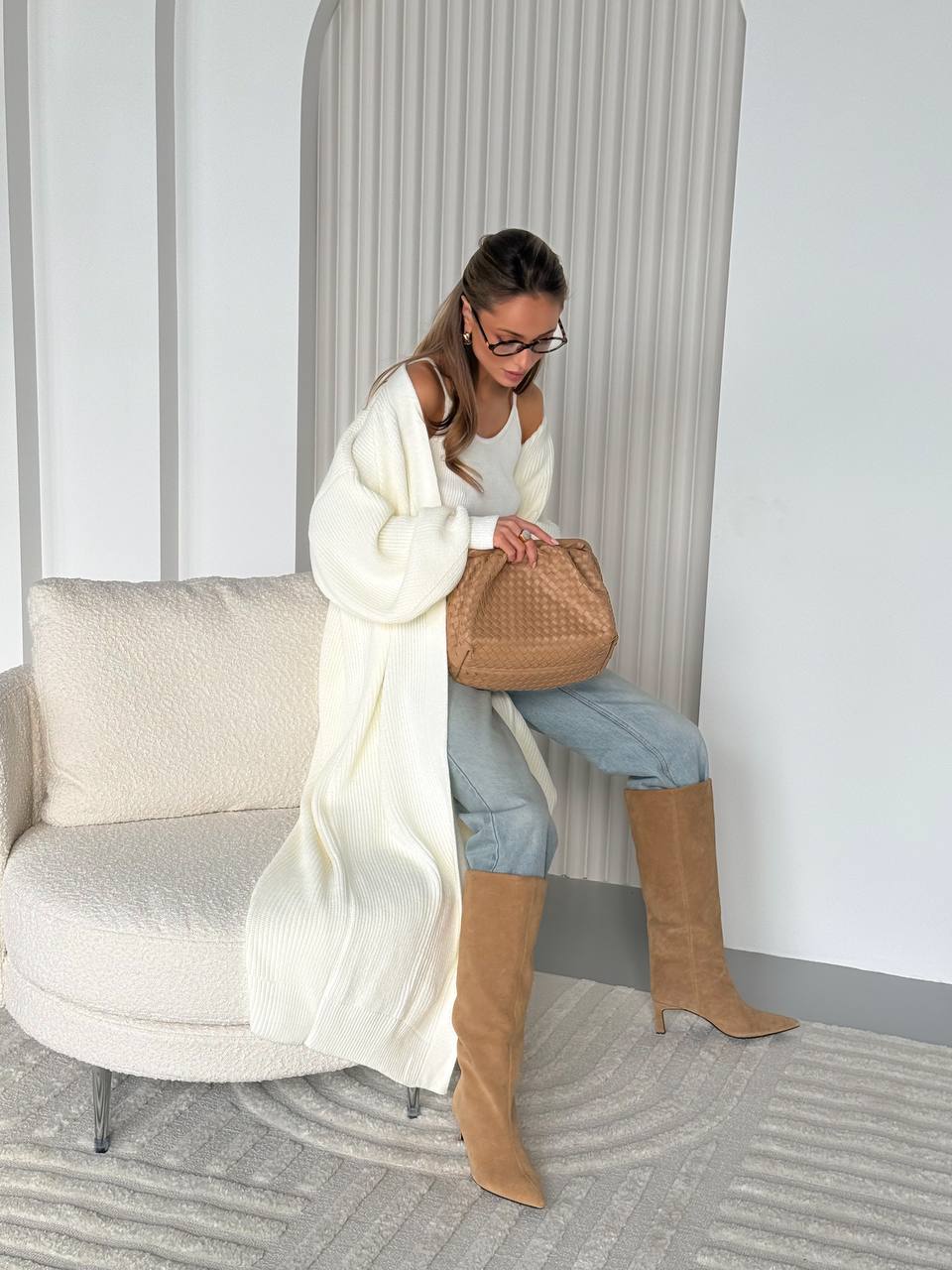 Woman in a white cardigan and tan boots sitting on a chair holding a brown bag.