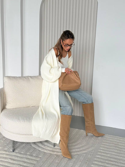 Woman in a white cardigan and tan boots sitting on a chair holding a brown bag.