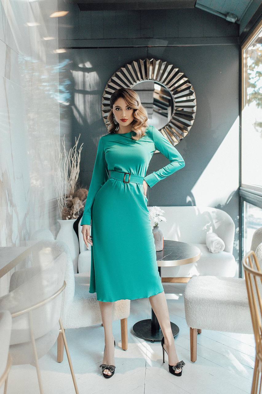 Woman in a teal dress standing in a modern interior setting with a decorative mirror.