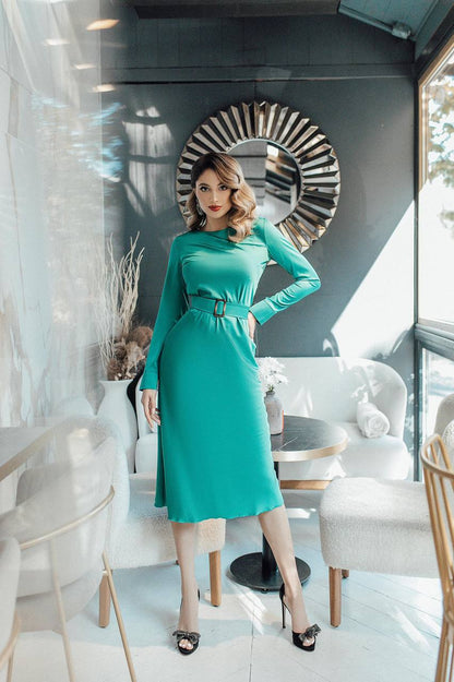 Woman in a teal dress standing in a modern interior setting with a decorative mirror.