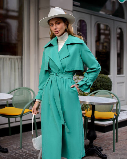 Woman in a teal trench coat and white hat standing outdoors near tables and chairs.