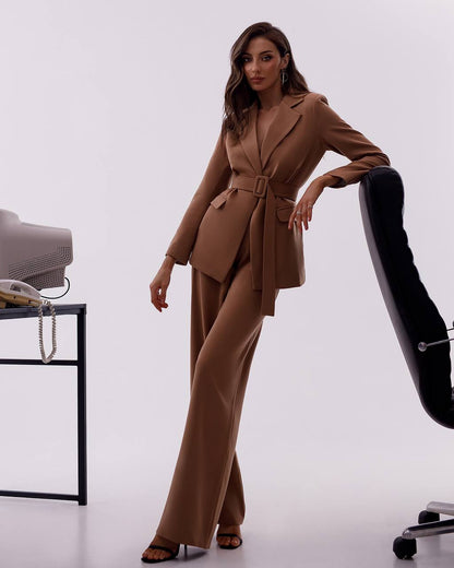 Woman in a brown suit standing in an office setting with a chair and desk.