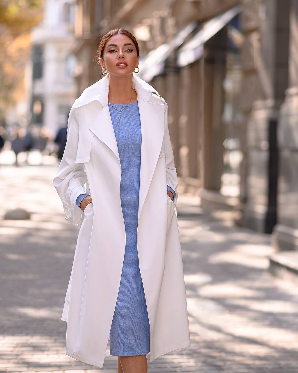 Woman wearing a white coat over a blue dress on a city street.