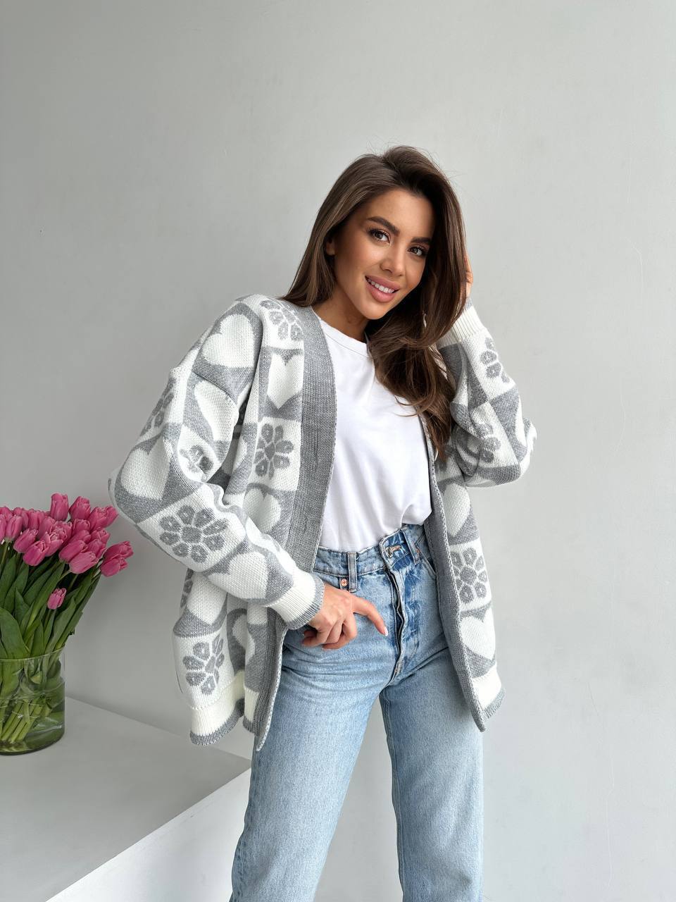 Woman wearing a patterned cardigan with a vase of pink tulips on a light gray background