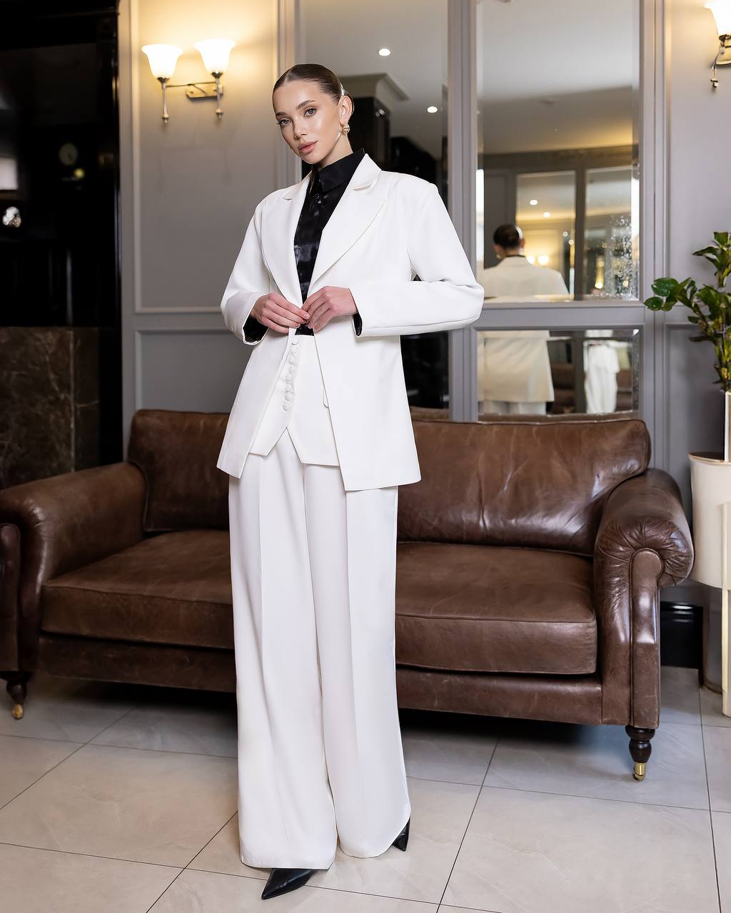 Woman in a white suit standing in a room with a brown leather couch and reflective surfaces.