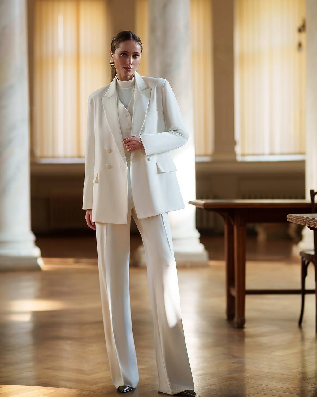 Woman in a white suit standing in a room with columns and wooden floors.