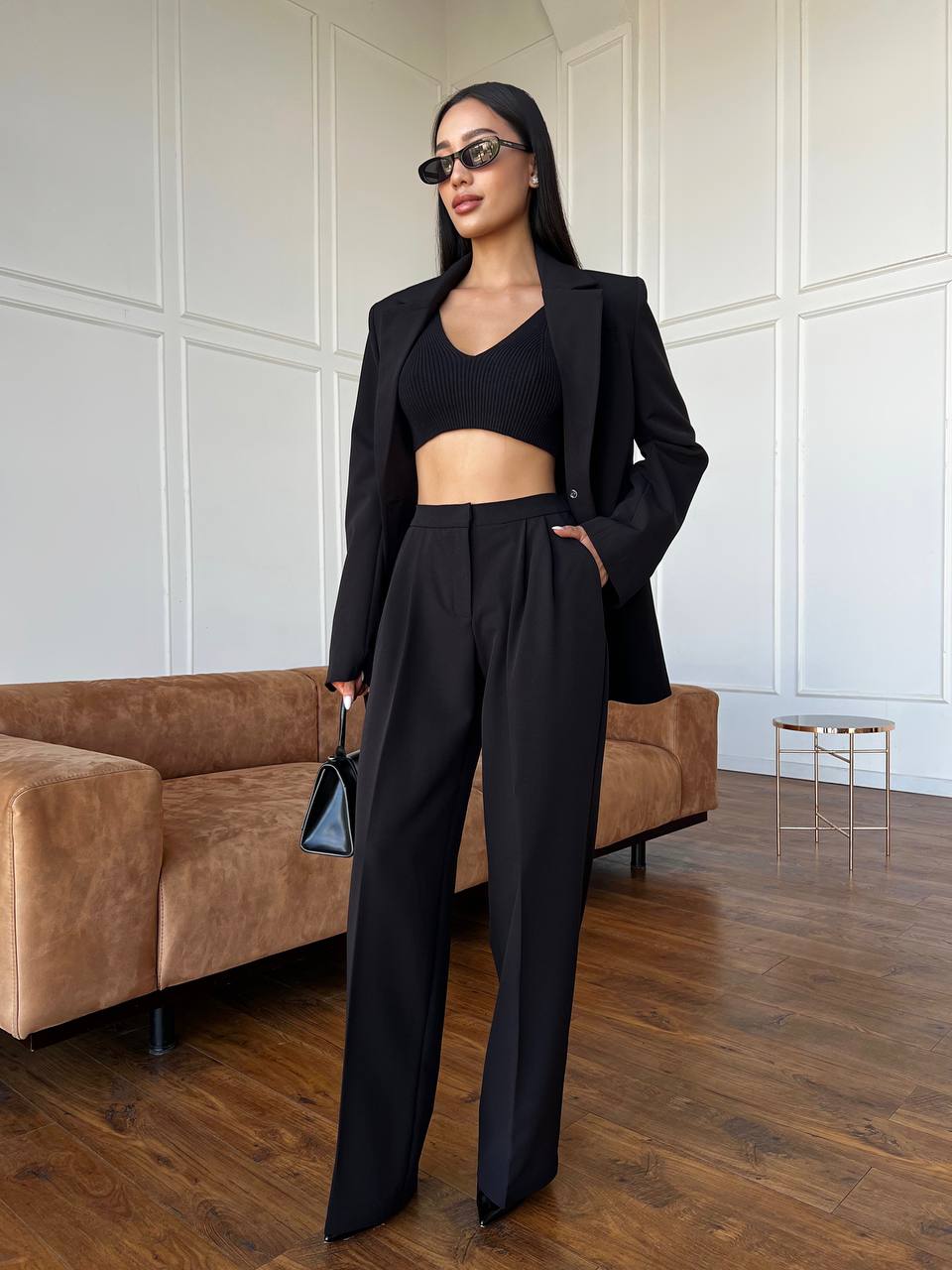 Woman in a black suit standing in a room with a brown couch and white walls.