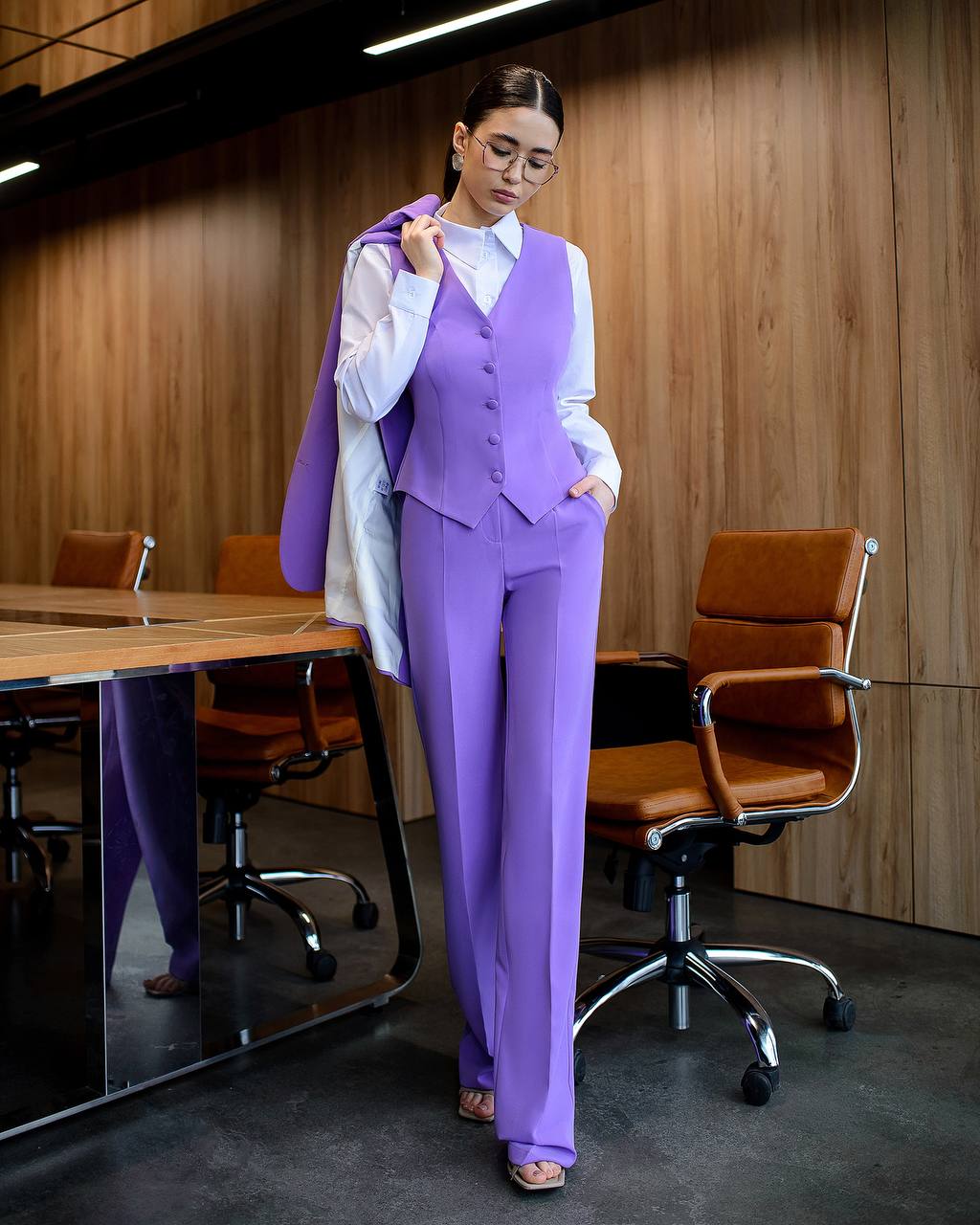 Woman in a purple suit standing in an office setting with wooden walls and a desk.