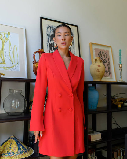 Woman wearing a red coat standing in front of a bookshelf with decorative items.
