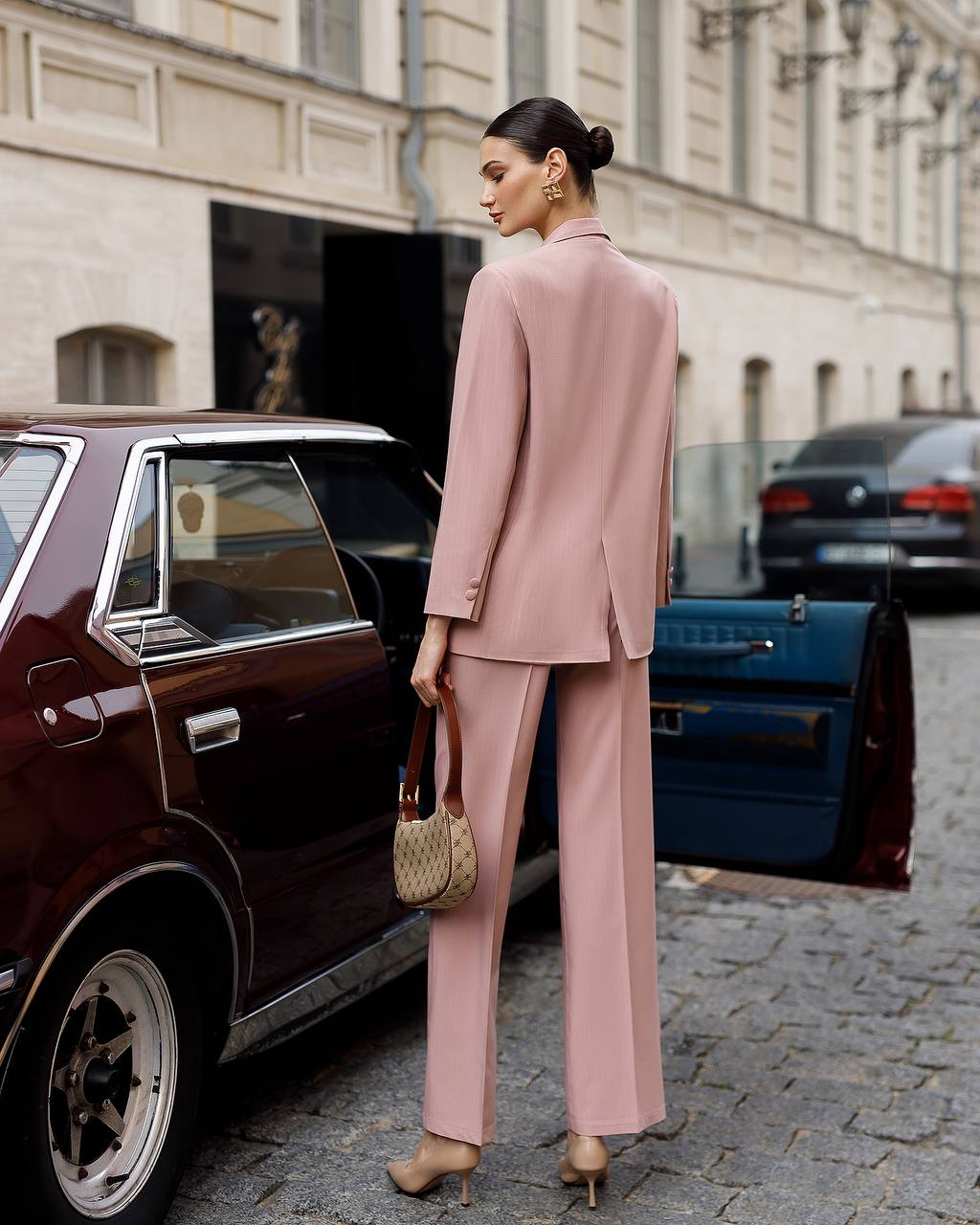 Woman in a pink suit standing next to a vintage car on a city street.