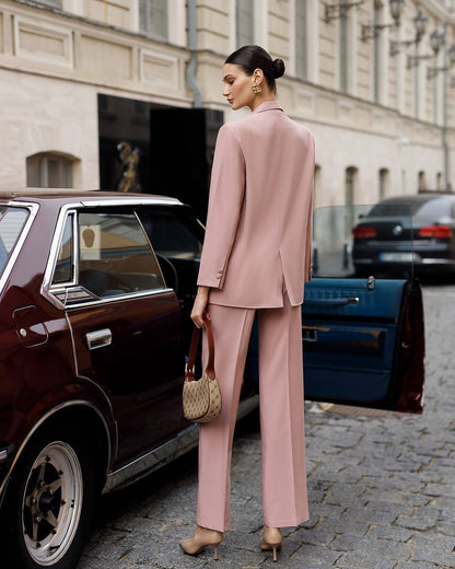 Woman in a pink suit standing next to a vintage car on a city street.