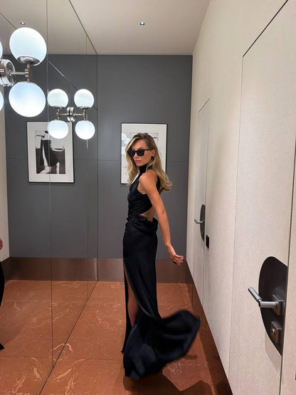 A woman wearing a black satin evening gown with side cutouts and a thigh-high slit, posing in a hallway with a mirror reflecting her profile.