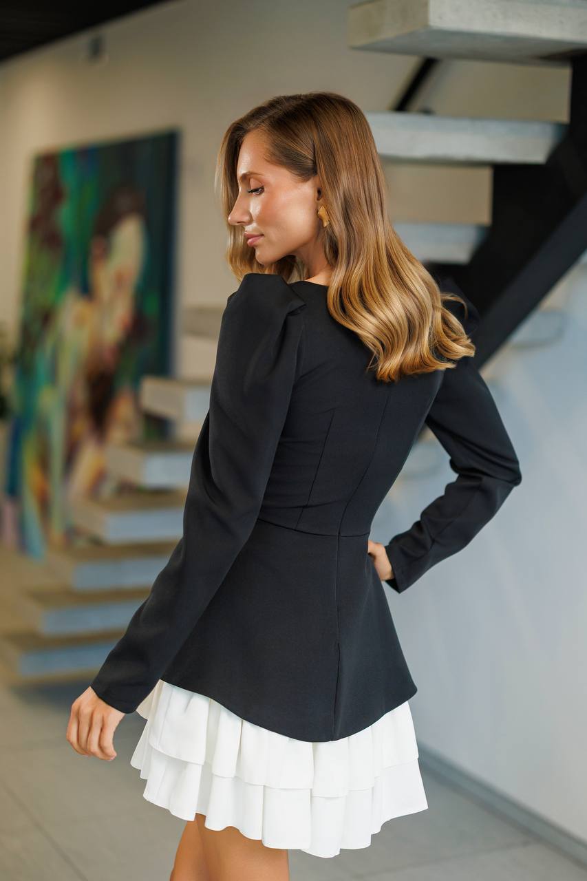 Woman wearing a black blazer and white skirt in an indoor setting with stairs and artwork in the background.