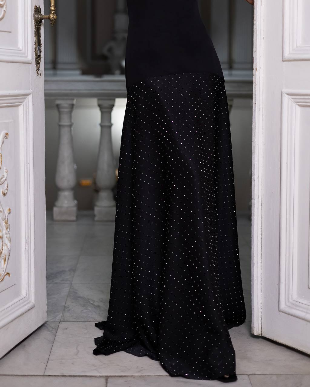 A black floor-length evening gown with a subtle dotted pattern, shown on a model standing in an open white doorway.