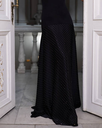 A black floor-length evening gown with a subtle dotted pattern, shown on a model standing in an open white doorway.