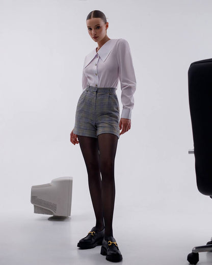 Woman in a light purple blouse and plaid shorts standing against a white background