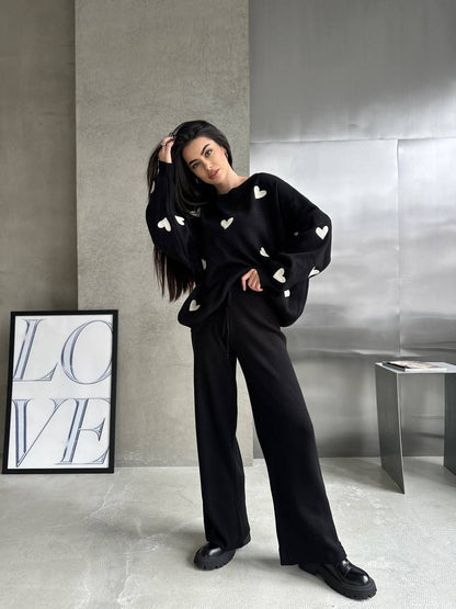 Person wearing a black sweater with white heart patterns and black pants in a modern interior setting.