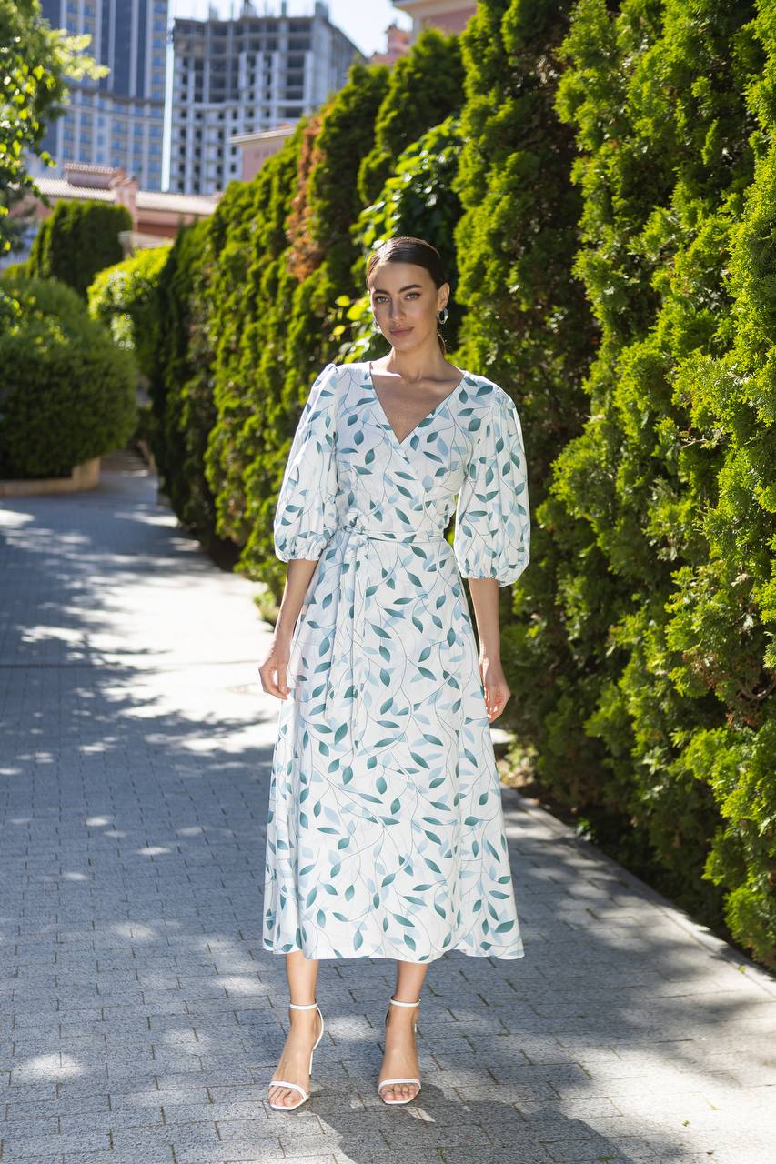Woman in a floral dress standing on a sidewalk with greenery and buildings in the background