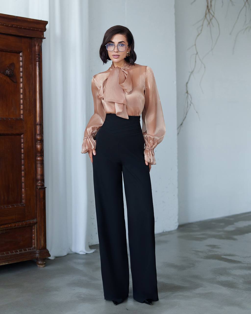 Woman wearing a sheer beige blouse and black pants in a minimalistic room.