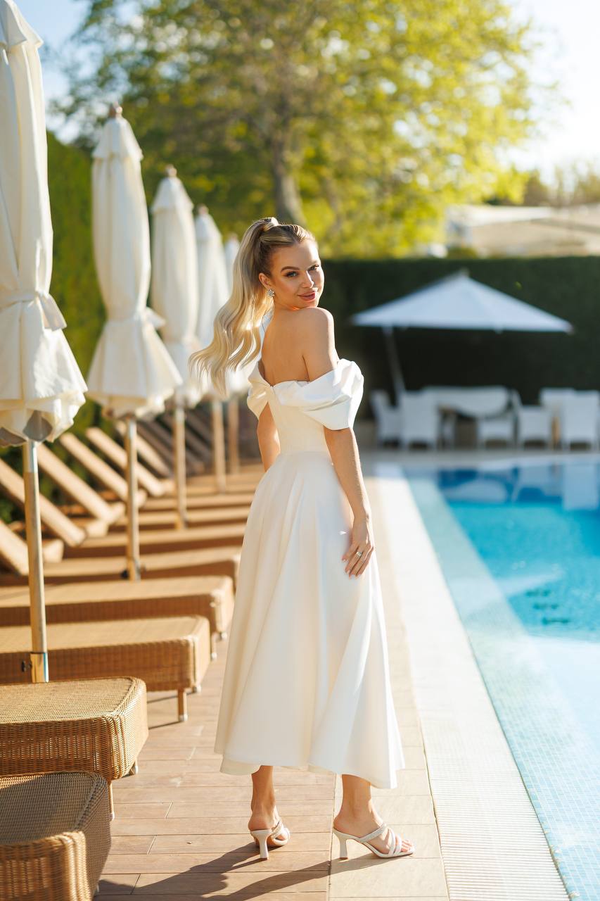 Woman in a white dress standing by a poolside with umbrellas and lounge chairs.