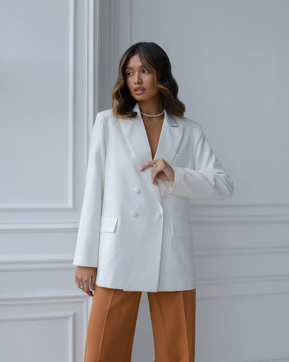 Woman wearing a white blazer and brown pants against a light gray wall.