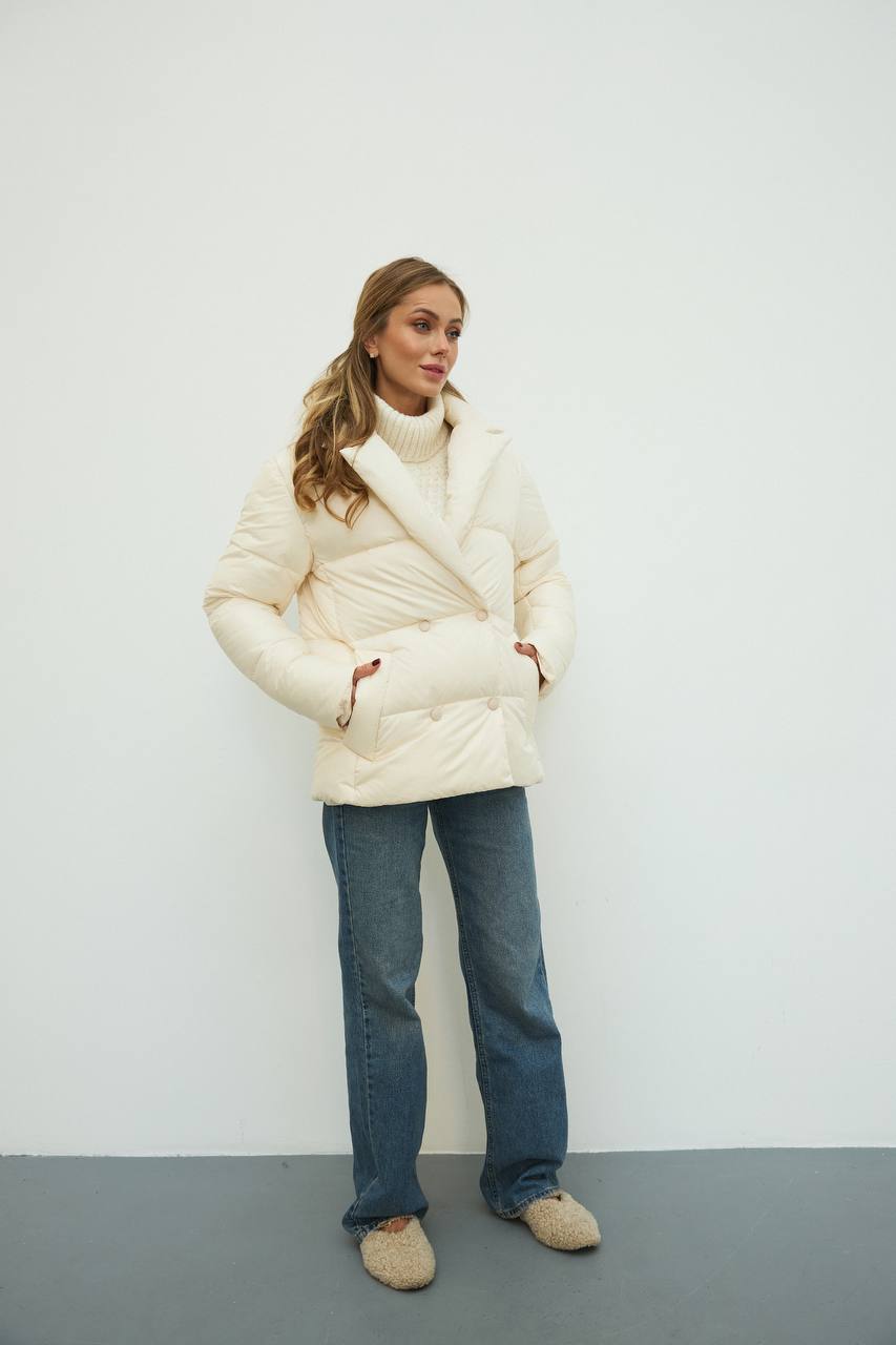 Woman wearing a white puffer jacket and blue jeans against a plain background