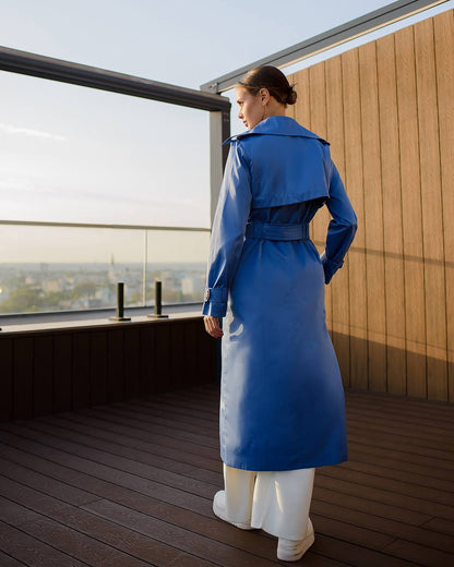 Woman in a blue trench coat standing on a rooftop with a cityscape view.