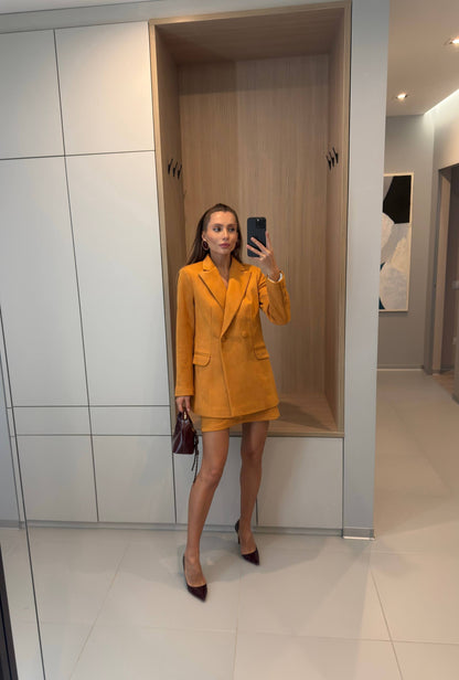 A woman takes a mirror selfie in a modern home interior, wearing a bright mustard yellow double-breasted blazer and matching mini skirt with dark pointed-toe heels.
