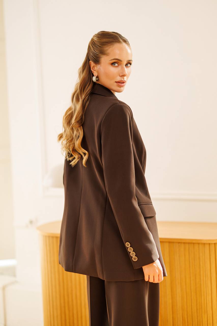 A woman with long wavy blonde hair is shown from the back wearing an oversized dark brown blazer and matching trousers in a bright indoor setting with a light wood cabinet.