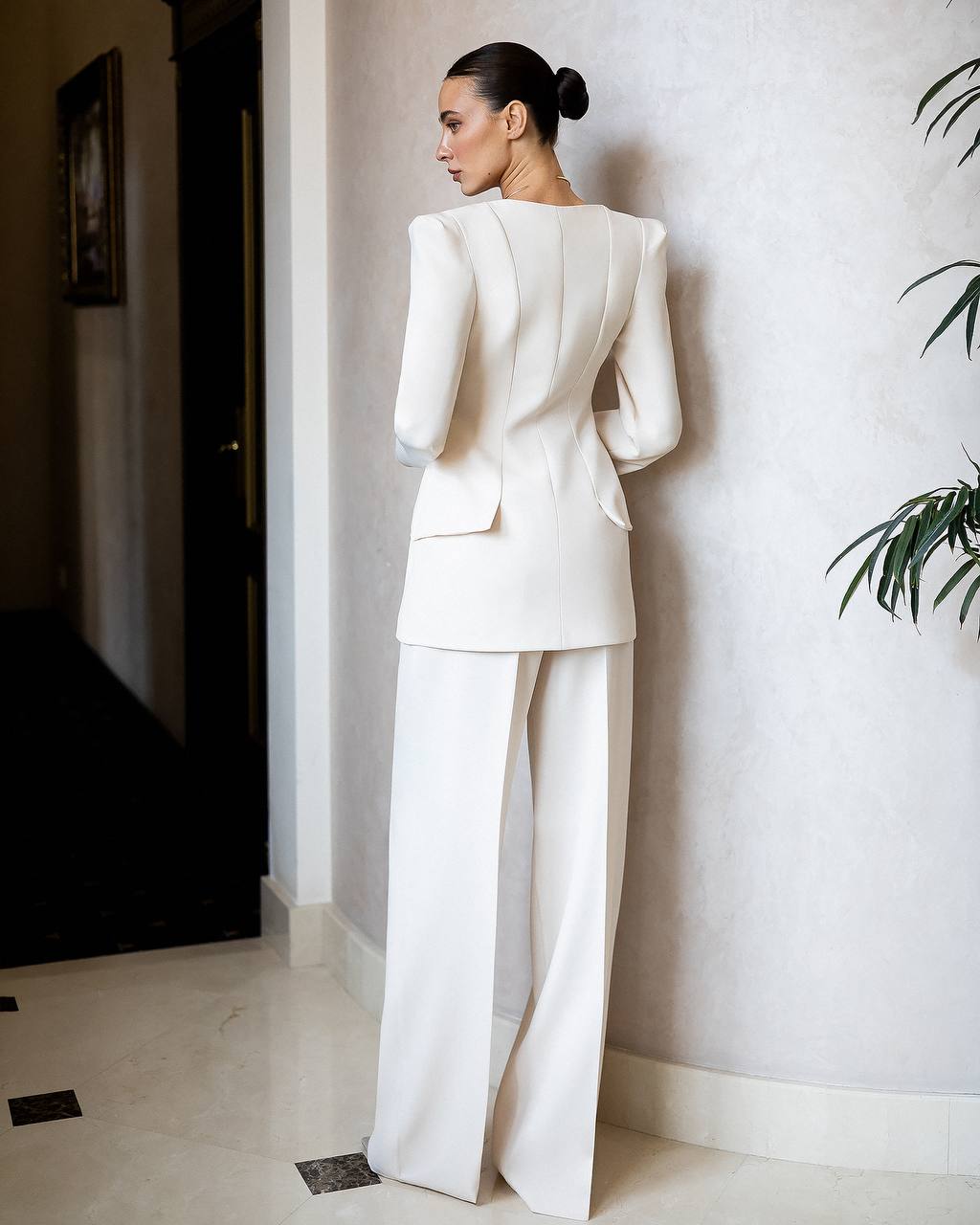 Woman in a white suit standing against a light-colored wall.