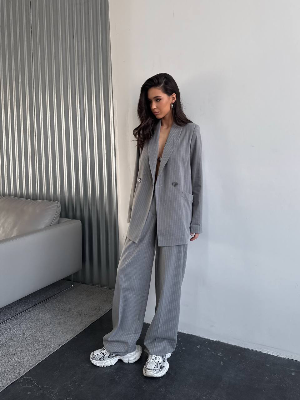 Woman in a gray suit standing in a minimalistic room with a bed and striped wall.