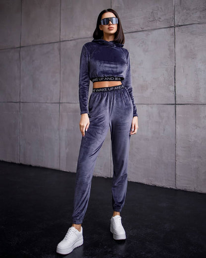 Woman wearing a gray velour tracksuit with text on it against a gray wall.