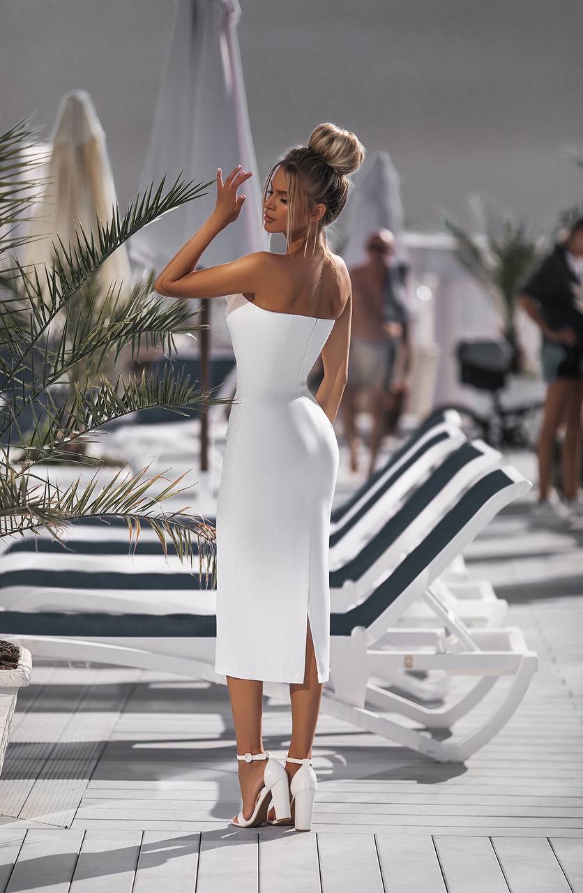 Woman in a white dress standing on a beach with lounge chairs and palm trees.